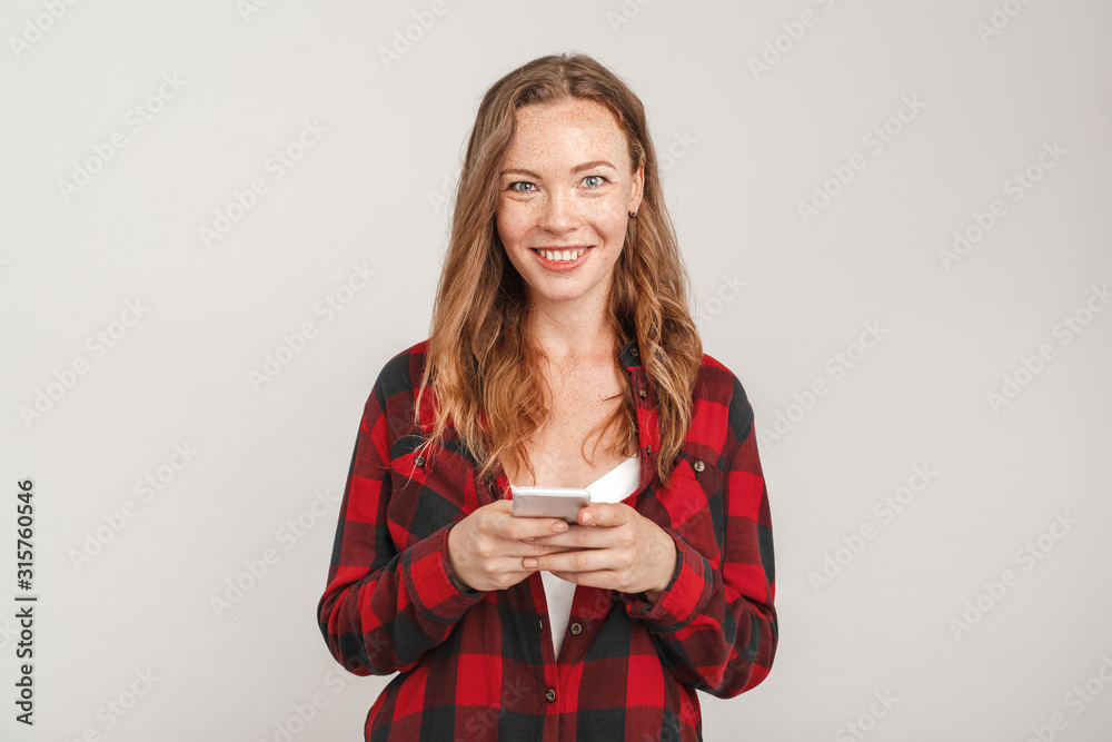 Freestyle. Young woman with freckles standing isolated on grey chatting on smartphone smiling excited Freestyle. Young woman with freckles standing isolated on grey chatting on smartphone smiling excited