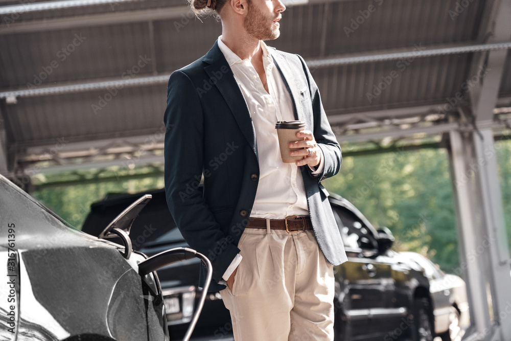 Mode of transport. Young stylish businessman in suit standing near electric car whiel charging drinking hot coffee Mode of transport. Young stylish businessman in suit standing near electric car whiel charging drinking hot coffee