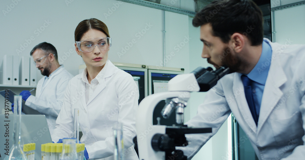 Obraz premium Portrait of the two Caucasian laboratory scientists, man and woman, talking while he working at the microscope and she at the laptop computer. Close up.