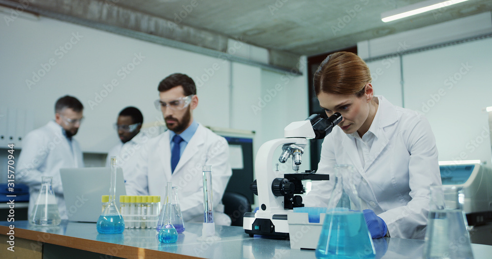 Caucasian woman scientist in the white robe looking in the microscope while doing some investigation and her male colleague testing some liquid in tube at the laptop computer. Caucasian woman scientist in the white robe looking in the microscope while doing some investigation and her male colleague testing some liquid in tube at the laptop computer.