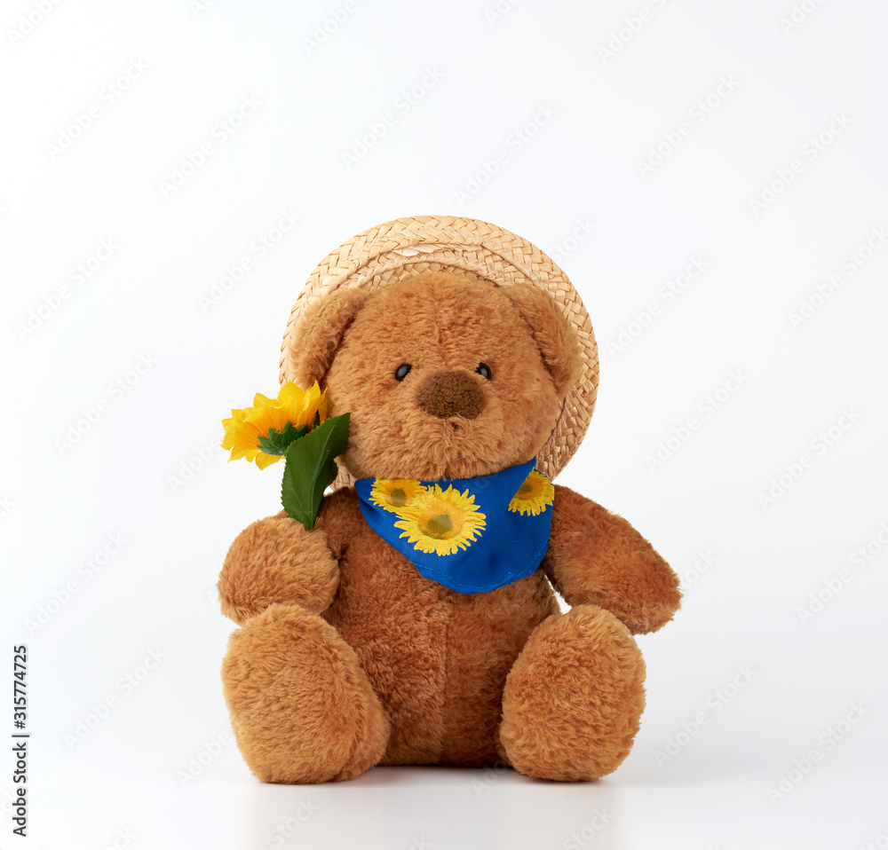 Obraz premium cute little brown teddy bear holds a plastic yellow sunflower and is dressed in a straw hat