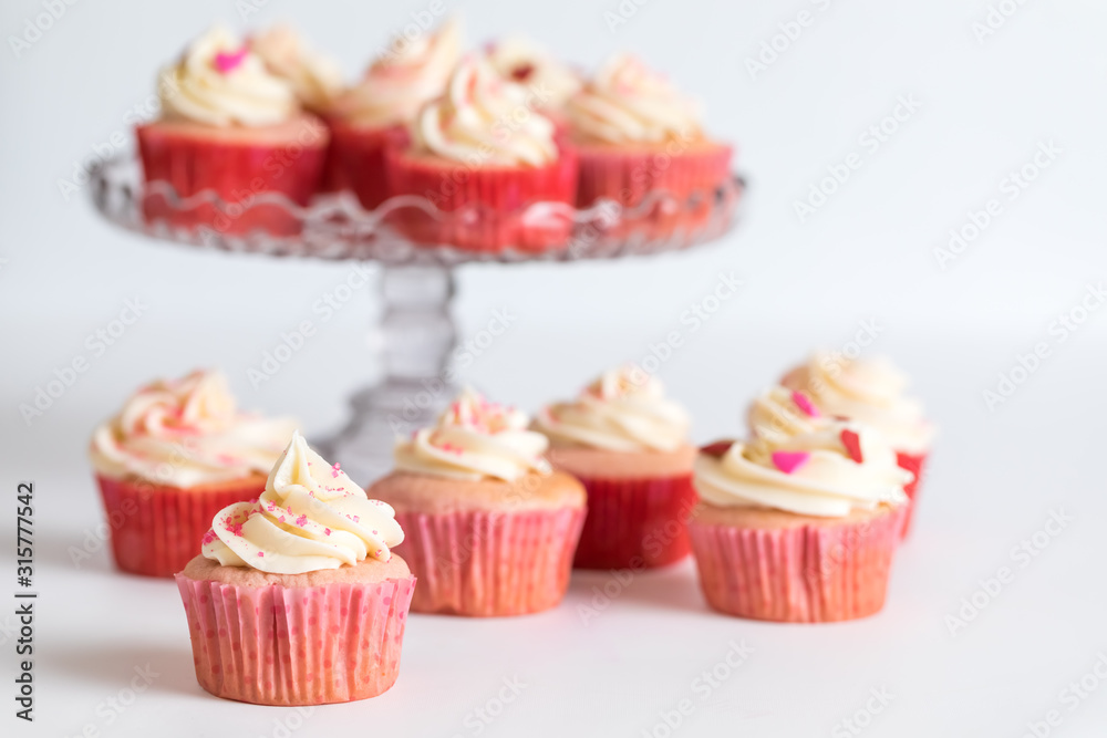 Strawberry cupcakes with cream cheese frosting on a cake stand with more on the table in front for Valentine's Day.