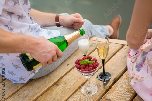 Romantic date surprise. A young guy and a girl on a wooden pier. The guy pours champagne into the glasses.