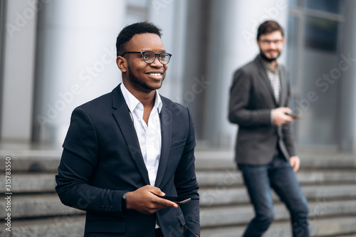 Young handsome african american businessman is walking on stairs smiling and holding a phone in his hand, outdoors