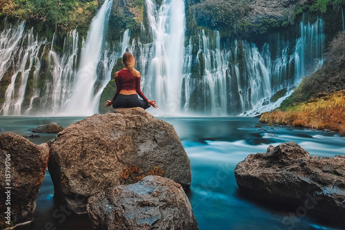 Woman meditating on top of a rock in front of a majestic blue waterfall