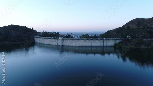 Aerial over blue water of Hollywood reservoir lake towards dam wall, Los Angeles city background