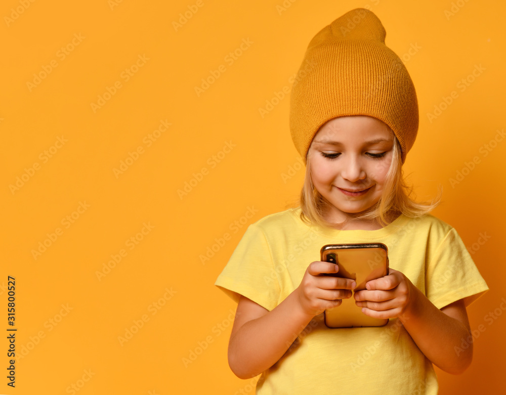 Little blonde kid dressed in t-shirt and hat, posing with smartphone against yellow background. Technology, children, internet. Close-up shot - obrazy, fototapety, plakaty 
