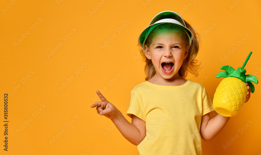 Little smiling girl in yellow t-shirt and hat holding fresh fruit juice in pineapple shaped bottle with straw and feeling fun Little smiling girl in yellow t-shirt and hat holding fresh fruit juice in pineapple shaped bottle with straw and feeling fun