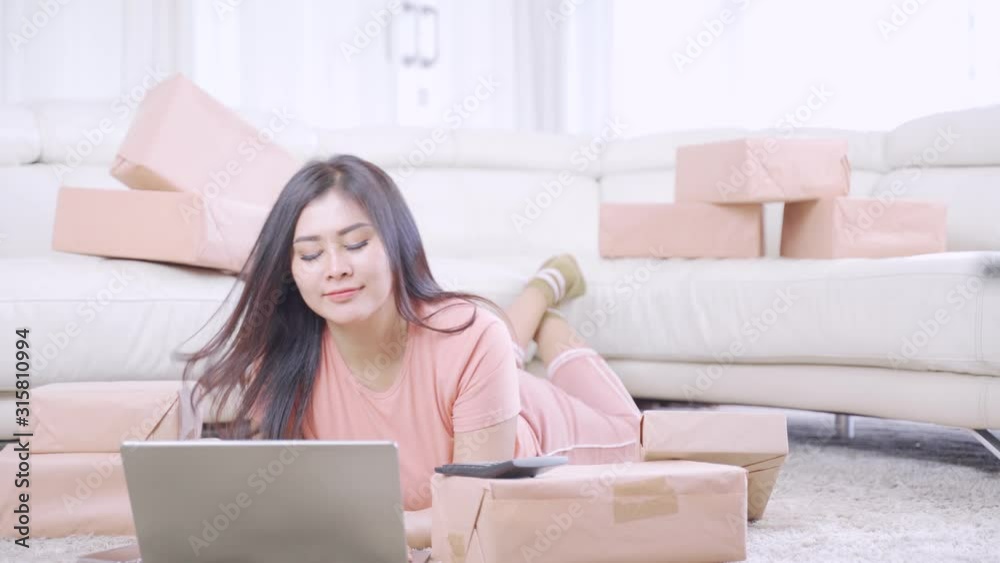 Beautiful woman online store owner working with laptop and cardboard while talking on mobile phone and lying on the carpet in the living room at home. Start up concept. Shot in 4k resolution