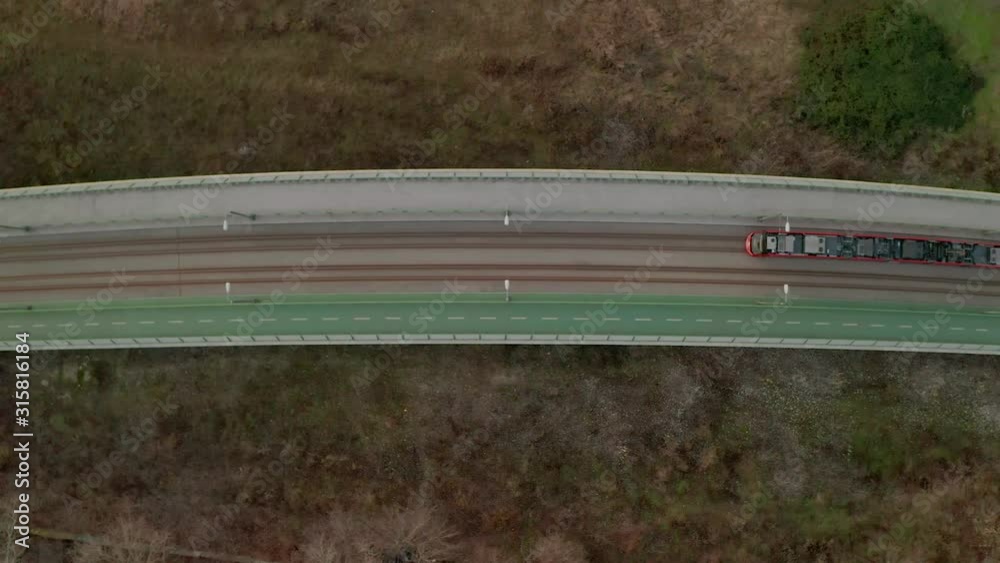 Bratislava tram driving on an elevated tram track above grass land. Aerial drone video with the camera tilted down, following the tram. Moving from right to left along a cycling path.