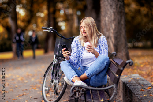Blonde young woman sitting on the bench looking on mobile phone and drinking coffee to go in park with bicycle on the background