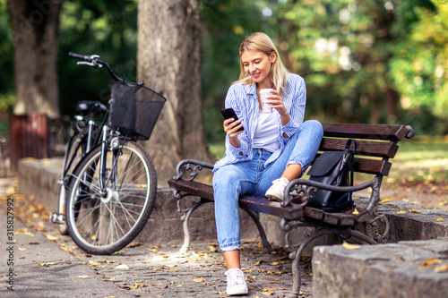 Blonde young woman sitting on the bench looking on mobile phone and drinking coffee to go in park with bicycle on the background