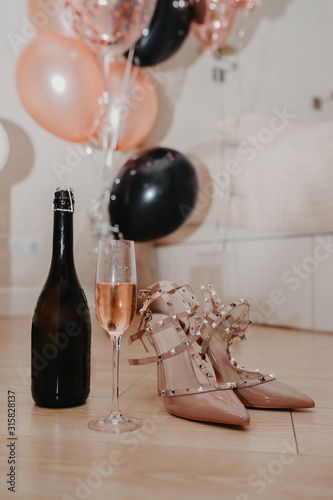 invitation card for a bachelorette party, stylish beige sandals with spikes, a glass of pink champagne and a bottle on a background of pink and black balloons horizontally