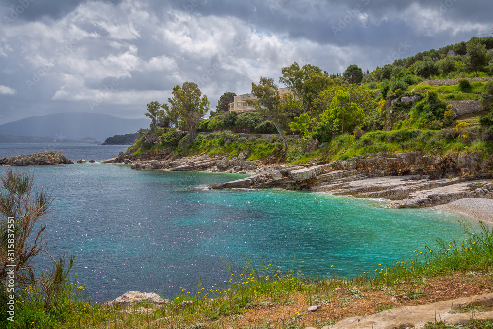 Fototapeta premium Beautiful landscape – sea lagoon with turquoise calm water, stones and rocks on the beach, cloudy sky and green trees. Corfu Island, Greece, Kassiopi village