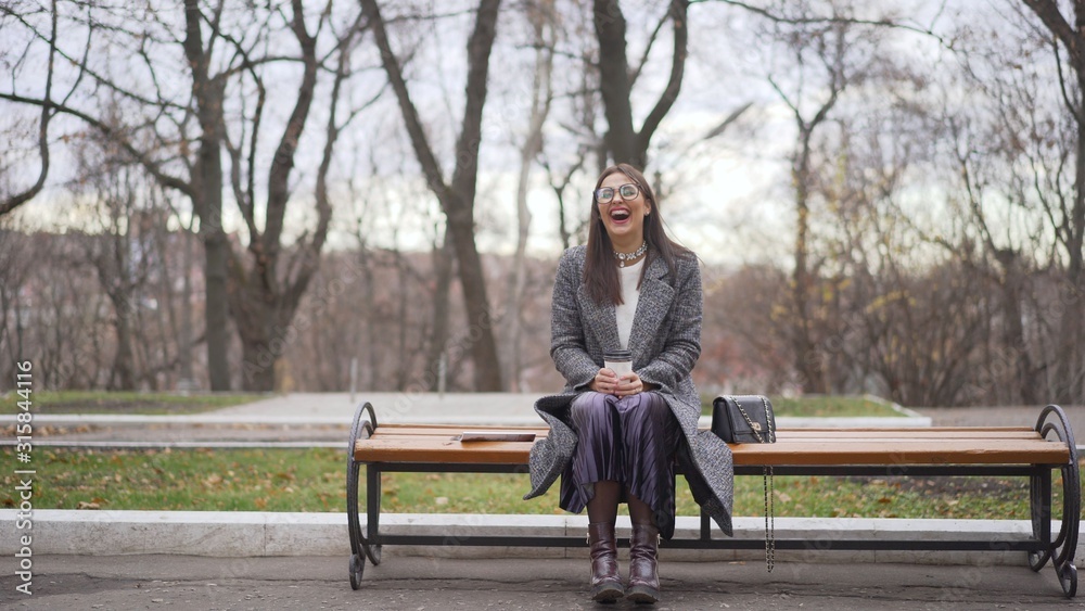 Fototapeta premium Portrait of a laughing girl dressed in an autumn coat holding a book sitting outdoors Girl laughs sitting on a bench in autumn.