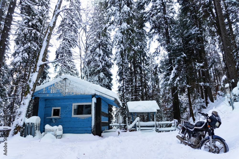 View at wooden buiding toned in classic blue in winter forest, motorcycle is on a side, snow is all around. Day time, good background. Winter forest concept. View at wooden buiding toned in classic blue in winter forest, motorcycle is on a side, snow is all around. Day time, good background. Winter forest concept.