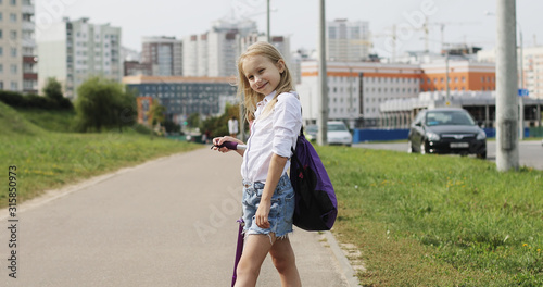 A cute blonde girl stands with a scooter on the streets along the road in the city. The child is resting in the summer.