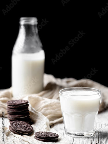 A glass with milk and several chocolate cookies on a wooden surface. Close-up. Bottle of milk in the background. Natural dairy products. Vertical frame, space for text. Shallow depth of field. photo