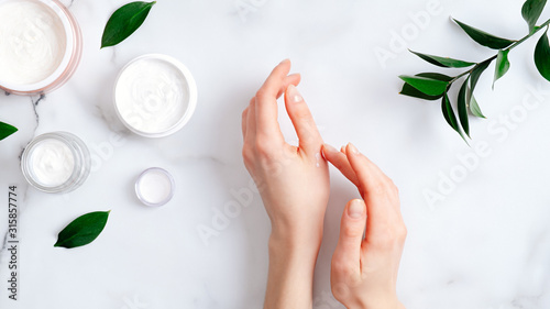 Cosmetic cream on female hands, jars with milk swirl cream and green leaves on white marble table. Flat lay, top view. Woman applying organic moisturizing hand cream. Hand skin care concept