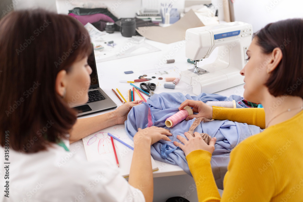 Two young female dressmakers working in sewing studio. Seamstress select color of threads suitable for fabric. Bespoke tailoring. Private enterprise. Hobby and business. Two young female dressmakers working in sewing studio. Seamstress select color of threads suitable for fabric. Bespoke tailoring. Private enterprise. Hobby and business.