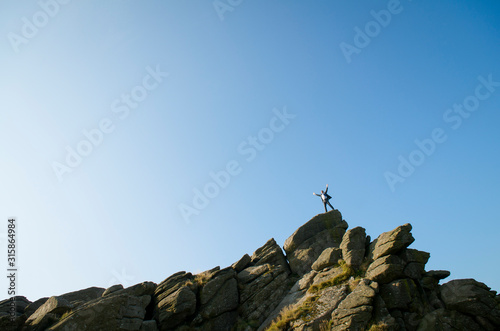 Distant businessman standing on the top of a rocky mountain peak celebrating in blue sky copy space