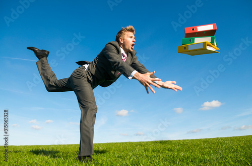 Clumsy businessman tripping dramatically with his file folders flying outdoors in a green grass meadow
