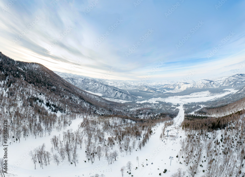 Aerial Picturesque panoramic landscape in the Altai mountains with snow-capped peaks under a blue sky with clouds in winter with gondola cableway and booths on ski resort. White snow and calm.