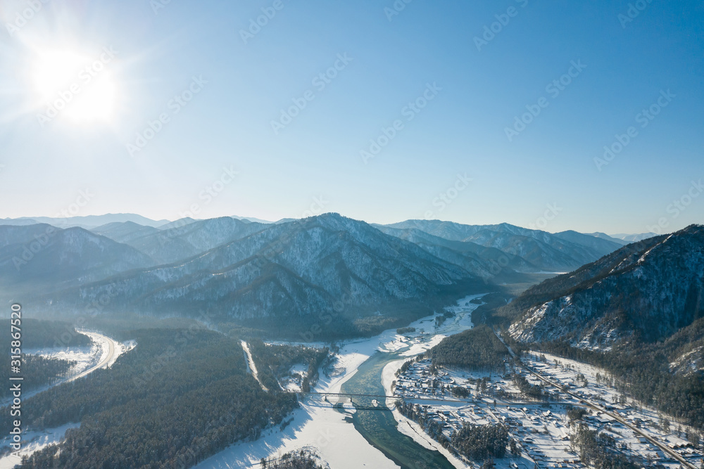 Picturesque landscape in the Altai mountains with snow-capped peaks under a blue sky with clouds in winter with green river and bridge for cars. White snow and calm.