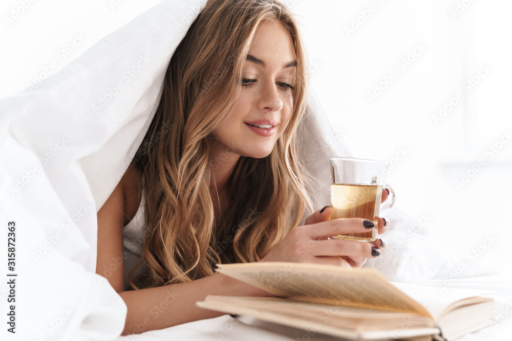 Photo of young focused woman reading book and drinking tea while lying Photo of young focused woman reading book and drinking tea while lying