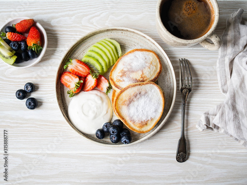 Delicious breakfast - fritters pancakes, sprinkled with powdered sugar with fruits and berries and sour cream on plate on wood table. Copy space. Top view