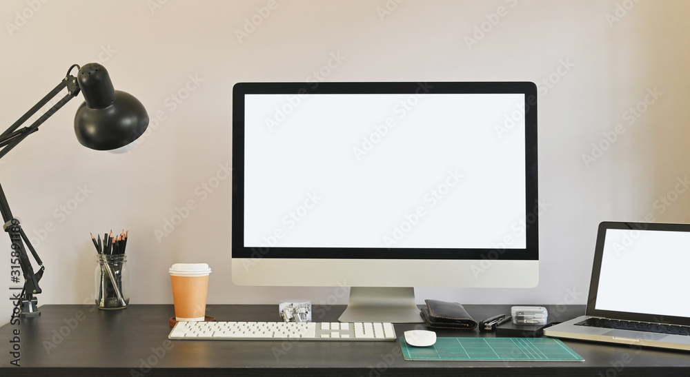 Fototapeta premium Photo of white blank screen computer and white blank screen laptop putting on the table together including lamp, coffee cup, mouse pencil holder and personal equipment. Modern working desk concept.