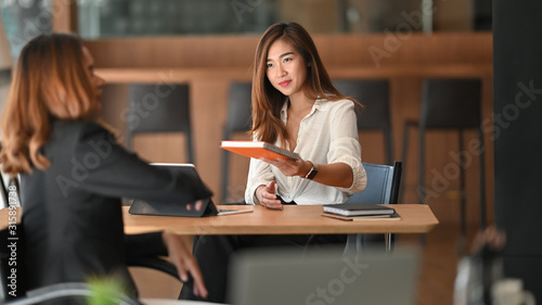 Young beautiful confident girl in white shirt giving a minutes of meeting to her colleague while sitting at wooden table in the modern office.