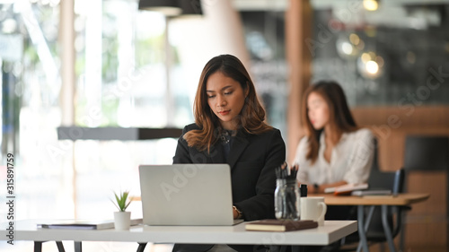 Photo of young business woman in look good black suit typing on her white laptop while sitting at work desk in the modern office. Sitting over colleague background.