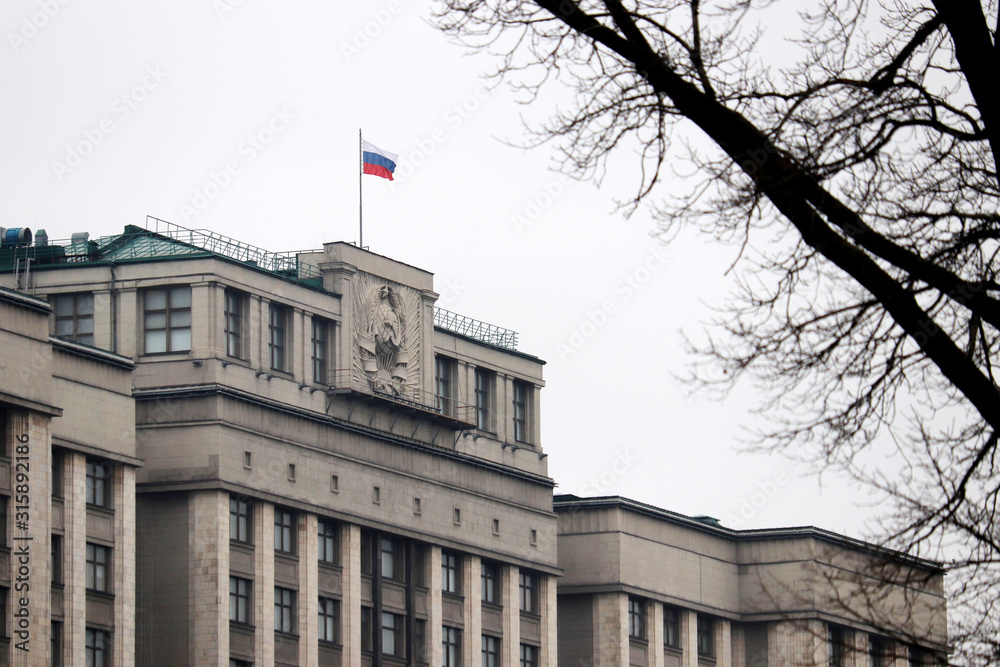 Obraz premium Parliament building in Moscow and bare tree branches against cloudy sky, selective focus. Russian authority, facade of State Duma of Russia with flag