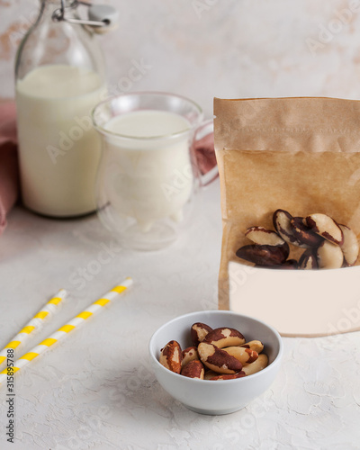 A handful of Brazil nuts in close-up  next to a package of nuts with space for text and logo. In the background  a mug and a bottle of milk.