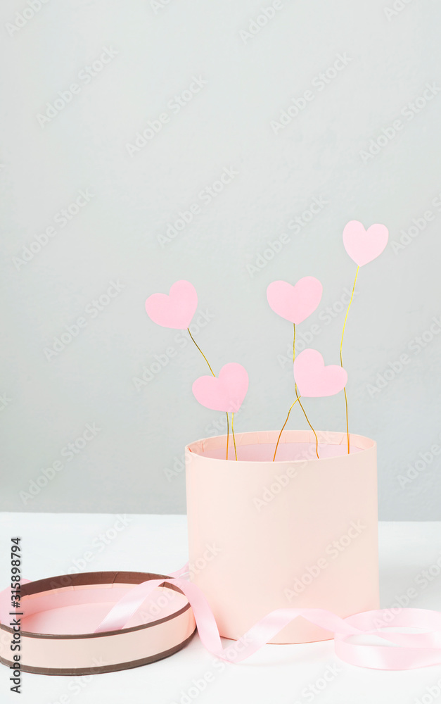 Round craft pink present box and sweet pink hearts in it on the white surface against grey wall.Vertical shot.Concept of valenite`s day and gifts