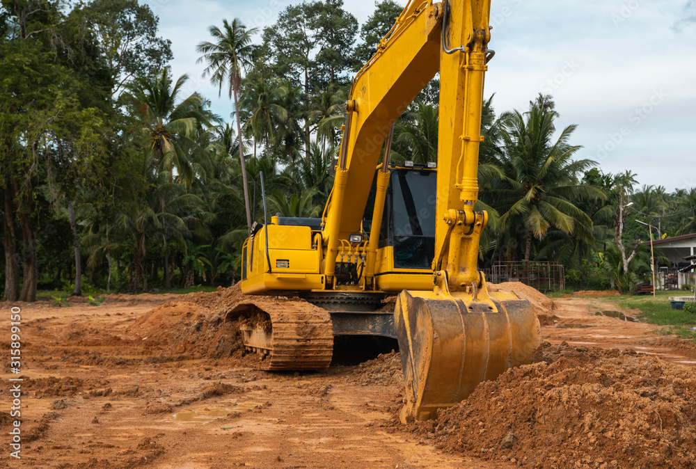 Yellow excavator on a construction site against blue sky. Heavy industry. Close up details of industrial excavator. Large tracked excavator standing on a orange ground with a palms on background. Yellow excavator on a construction site against blue sky. Heavy industry. Close up details of industrial excavator. Large tracked excavator standing on a orange ground with a palms on background.