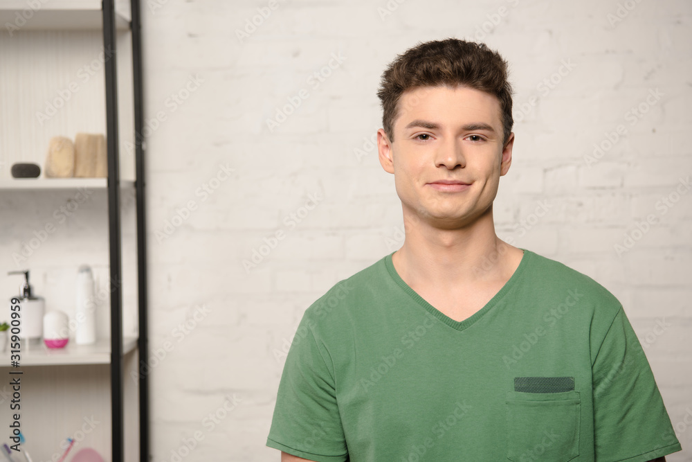 Obraz premium handsome young man in green t-shirt smiling at camera while standing near rack with cosmetics