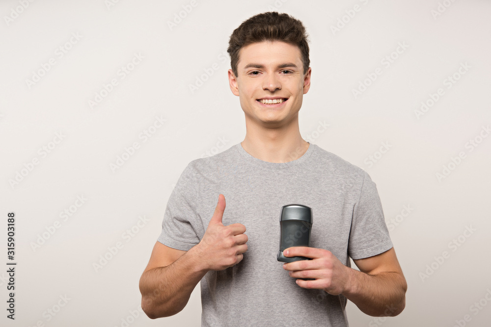 smiling man in grey t-shirt holding deodorant and showing thumb up isolated on grey smiling man in grey t-shirt holding deodorant and showing thumb up isolated on grey