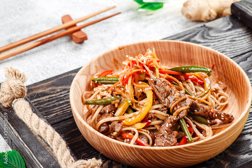 Pan-Asian food. Noodles with vegetables and meat in a beautiful wooden plate on a dark rustic tray on a light concrete background. Beautiful composition. Close-up. Space
