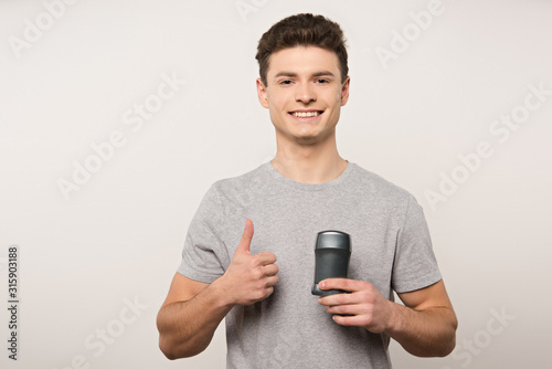 smiling man in grey t-shirt holding deodorant and showing thumb up isolated on grey