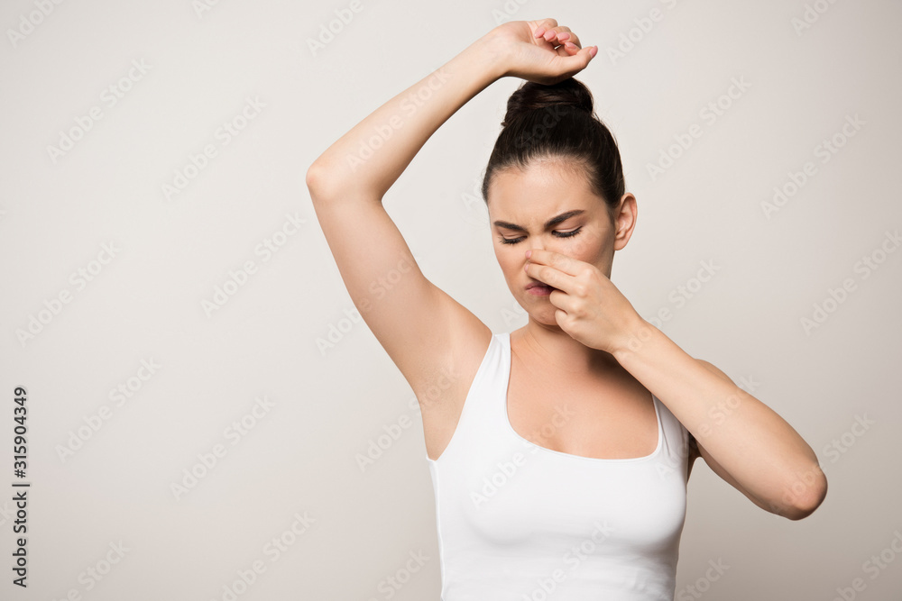 displeased woman plugging nose with hand while looking at underarm isolated on grey displeased woman plugging nose with hand while looking at underarm isolated on grey