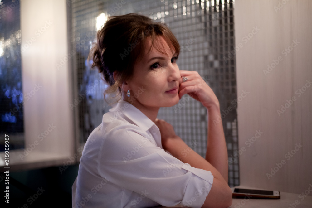 Adult beautiful woman sit in restaurant and waiting the order in evening. The female person wearing white shirt. Adult beautiful woman sit in restaurant and waiting the order in evening. The female person wearing white shirt.