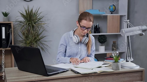 Close view of satisfied pretty young lady in casual clothes wearing glasses which writing notes from book into notebook photo