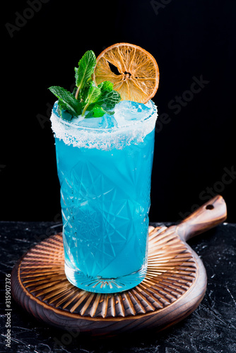 Blue cocktail with ice, decorated with mint and slice of dry orange in beautiful faceted glassm on black background photo