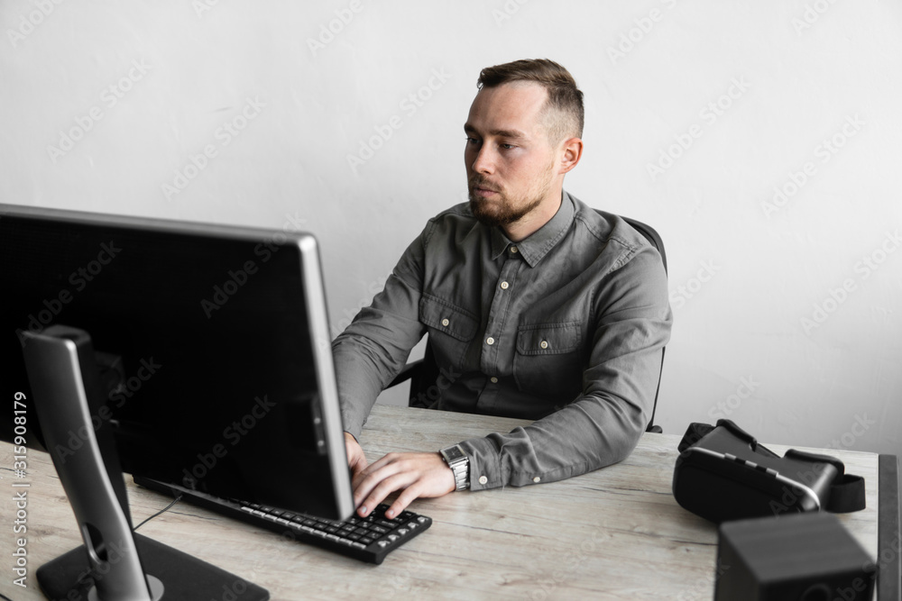 Young businessman or student in a shirt sitting against monitor of computer. Working on a pc at a table in the office with a thoughtful expression. Young businessman working on his laptop in office. Young businessman or student in a shirt sitting against monitor of computer. Working on a pc at a table in the office with a thoughtful expression. Young businessman working on his laptop in office.