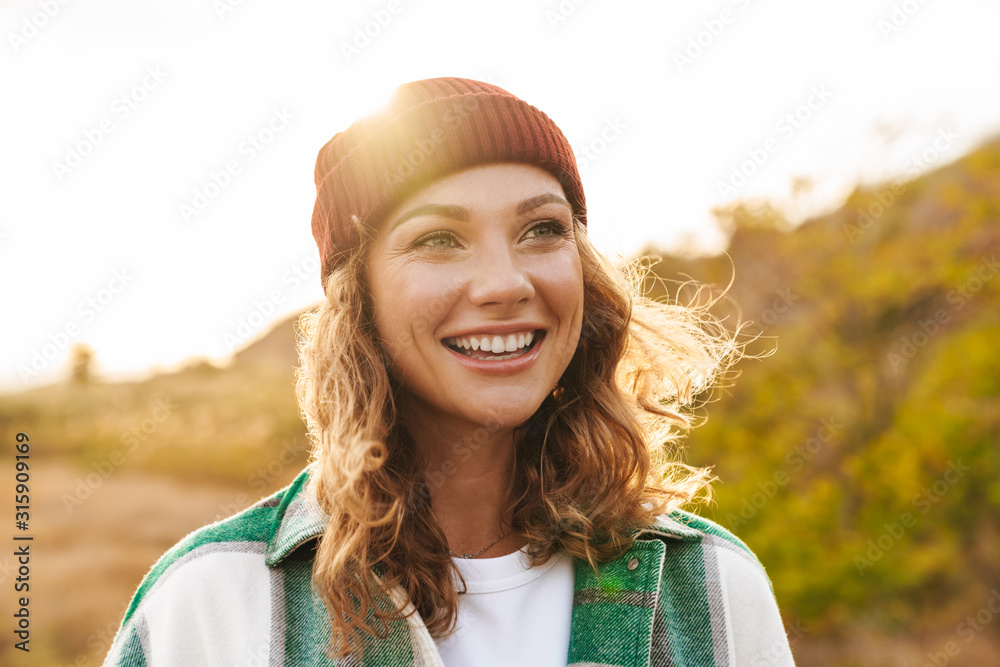Image of young woman wearing hat and plaid shirt walking outdoors - obrazy, fototapety, plakaty 