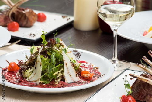 Fresh salad with beef carpaccio, parmesan, tomatoes and sauce on a white plate and a glass of white wine on a table in a restaurant. Close-up. Space