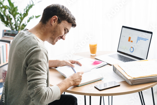 Image of young man doing calculations while working on laptop at home