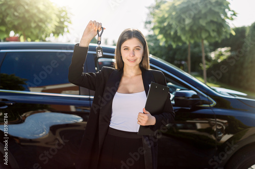 Beautiful girl with car key in hand. Caucasian woman car seller holding car keys, standing in front of new black car outdoors in vehicle trade fair. Auto rental or sales concept. © sofiko14
